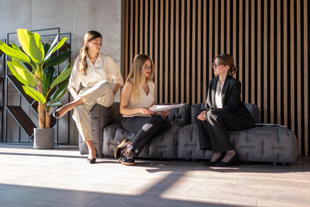Three businesswomen are sitting on a modern office sofa, discussing important partnership details...