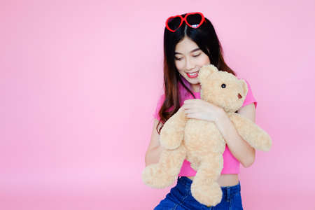 Portrait beautiful asian cheerful girl with red heart shaped sunglasses and beaming smile on pink background with teddy bear holding in hand, emotions action, happy summer lifeの写真素材