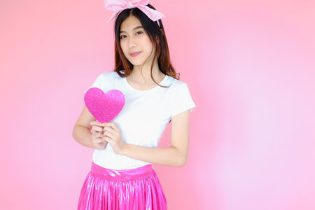 Portrait beautiful asian teen girl wearing white T-shirt and pink skirt on pink background, happy valentine day in love concept, model holding red heart sign in handの写真素材