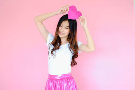 Portrait beautiful asian teen girl wearing white T-shirt and pink skirt on pink background, happy valentine day in love concept, model holding red heart sign in handの写真素材
