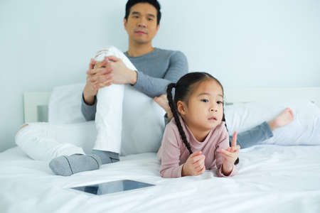 Single dad teaching daughter using a tablet on bed in bedroom, father tired conceptの写真素材