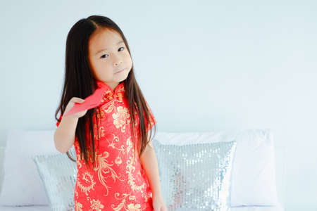 Portrait beautiful cute asian child girl wearing red mandarin gown or cheongsam dress on the bed with red envelopes holding in hand, celebrate chinese new year conceptの写真素材