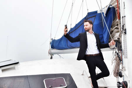 Businessman on luxury yacht with smartphone, handsome man wearing white shirt and suit on sailing boatの写真素材