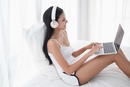 Asian woman sitting on the bed in morning with headphone and laptopの写真素材