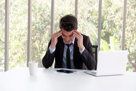 business caucasian man headache and sitting in working with laptop in office near window
