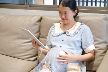 pregnant woman listening to music on belly use tablet with earphones on sofa at home in living roomの写真素材