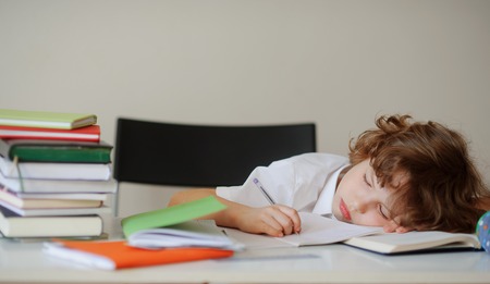 Child fell asleep while doing homework. On the table, textbooks and notebooks. In his hand the boy ballpoint pen. He put his curly head on an open book.の写真素材