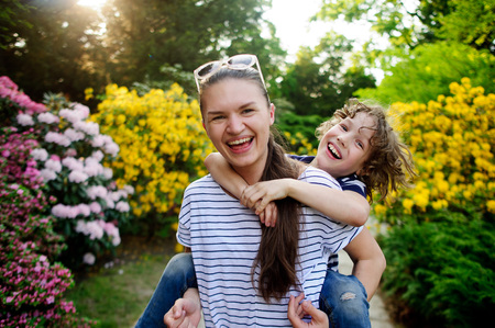 Young woman carries on her shoulders disheveled boy. They laugh. Behind them are picturesque flowering shrub.の写真素材