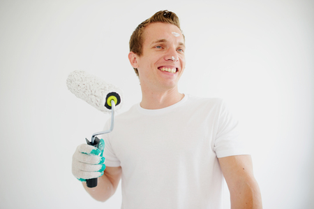 The young man holds in hand the roller for coloring and smiles. His face is soiled by white paint. The guy to stand. He looks aside. Behind the back there is a white wall.の写真素材