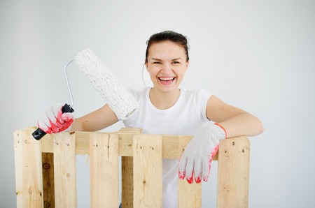 The cheerful young woman leans against a wooden pallet. Her hands in working gloves. She holds the roller for coloring. The woman looks in a chamber and laughs. On a white background of a wall.の写真素材
