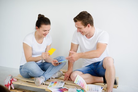 Young people select color for coloring of walls in the room. The guy and the girl sit on a floor in the empty room. They examine samples. On faces of a smile. Near them working gloves and catalogs.の写真素材