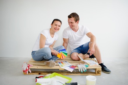 The young couple selects color of a decor in the new apartment.They sit on a floor on the wooden pallet and smile. The girl looks in a camera. The guy on the girl. White background of a wallの写真素材