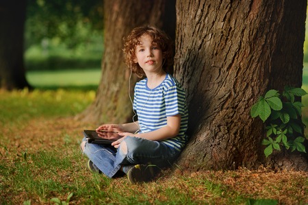 The boy of 8-9 years sits in park under a tree with the tablet in hands. The little fellow with a blond curly hair looks in a camera. In ears earphones. Behind the back there is a thick tree trunk.の写真素材