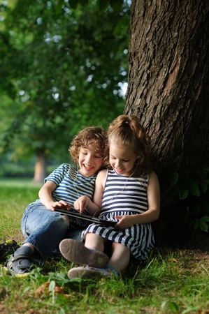 The boy and the girl of 7-8 years smiling look in the tablet. Children sit leaning backs against a trunk of a huge tree. They with interest look in the tablet screen. In their ears earphones.の写真素材