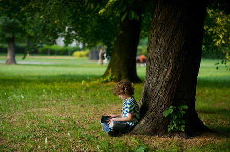 The little boy with the tablet on a lap sits under a huge tree. The boy in jeans full of holes sits having crossed legs. He thoughtfully looks at the screen.The boy is sideways to the camera.の写真素材
