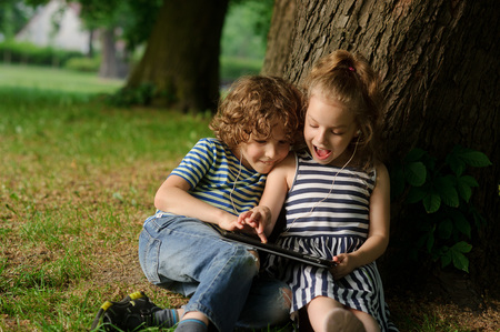 Brother with sister of 7-9 years sit under a tree and look in the laptop.Children sit having leaned against a trunk of a old tree and with enthusiasm look at the screen.They have one earphones for twoの写真素材