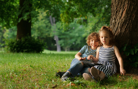 The boy and the girl of 7-8 years sit under a big tree in park.Children sit on the earth having extended legs.They hold the small laptop.On a background green park.Children look in a camera.の写真素材