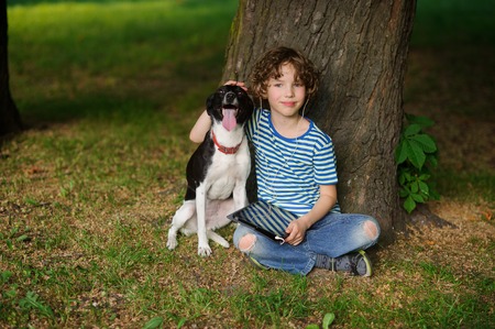 The boy with a dog sits on the earth under a tree. The child sits having crossed legs. He has put a hand on the head of a pet. On a lap the boy has a tablet. On a face a thoughtful smile.の写真素材
