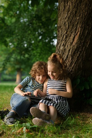 Two children sit in park under a big tree and play on the tablet. . They sit having leaned against a trunk of a big tree. On a lap tablet. In ears one earphones for two.の写真素材