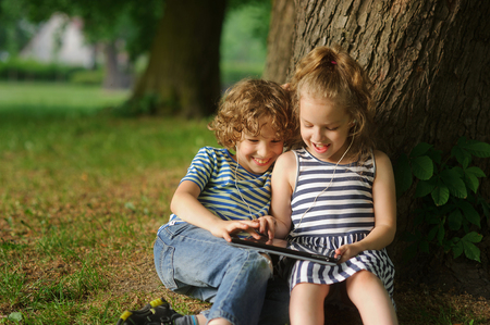Two children sit in park under a big tree and play on the tablet. The boy and the girl are keen on game. Him it is very cheerful.の写真素材