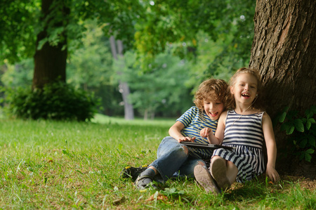The boy and the girl of 7-8 years sit under a tree having nestled and play on the tablet. Children are very keen. Them that - that has made laugh. Children laugh.の写真素材
