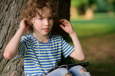The boy with the tablet listens to music. The blue-eyed curly boy in earphones has leaned against a...