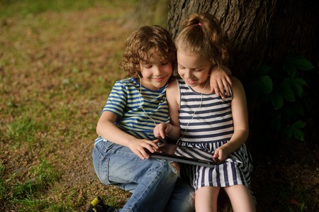 Two children sit under more tree with interest looking at the tablet screen. The boy and the girl sit having embraced. They smile, having inclined over the tablet.の写真素材