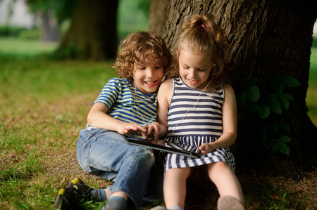 The brother and the sister of 8-9 years play with passion on the tablet. They sit having leaned against a trunk of a big tree. On a lap tablet. In ears one earphones for two.の写真素材
