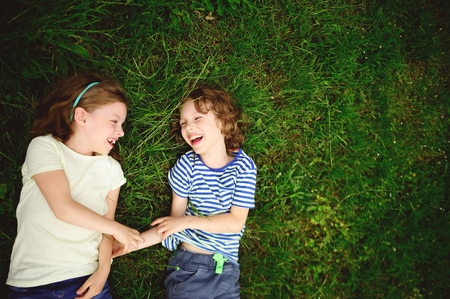 Two cheerful children lie on a green grass. The boy and the girl lie having joined hands and laugh. They look at each other.の写真素材