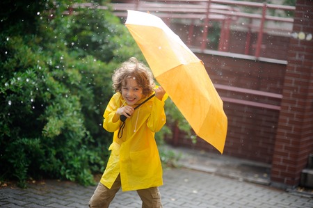 Boy of 8-9 years in the rain. It holds a yellow umbrella. The child with a smile looks in the camera having squinted. It has wild curly hair and a nice face. The boy is dressed in yellow raincoat.の写真素材