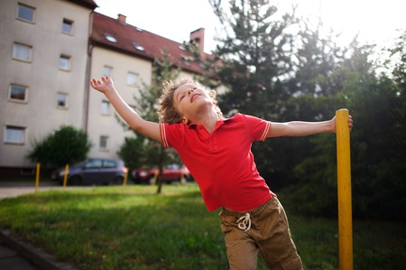 The little boy of 8-9 years turns around a support in the yard of the house. He imagines something during game. The boy has thrown back the head and has closed eyes from delight.の写真素材