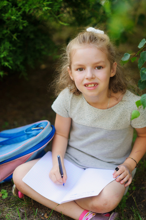 Cute schoolgirl sits having crossed legs under a tree and holds on a lap an open notebook. It has in a hand a ball pen. Little girl with a smile looks in the camera. Near the girl her satchel lies.の写真素材