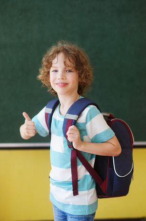 The cheerful pupil stands near a board in a class. He with a smile looks in the camera. Behind the back of the school student a backpack. Gesture of the boy shows that at it everything is all right.の写真素材