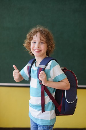 The cheerful pupil stands near a board in a class. He with a smile looks in the camera. Behind the back of the school student a backpack. Gesture of the boy shows that at it everything is all right.の写真素材
