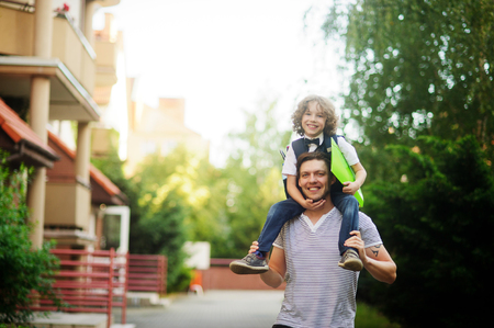 The father carries on his shoulders of his son-schooboy. Little learner with one arm hugs dad for the neck and the other holding a green folder. The boy's curly blond hair and a nice face. Father and son smile.の写真素材