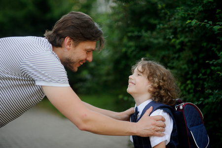 The father sees off the son-schoolboy in school. It has bent to the boy and has taken it for shoulders. The father and the son look against each other with love and smile.の写真素材