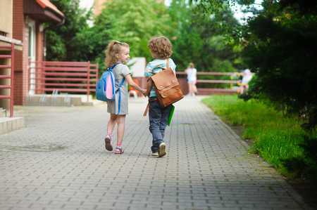 Young students, boy and girl, going to school. They hold hands. They hold hands. Children behind shoulders have satchels. Warm day in an early autumn. Back to school.の写真素材