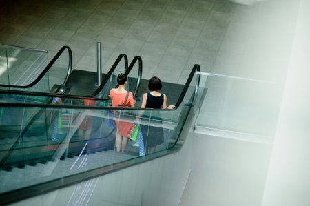 Two young darkhaired woman on the escalator at the mall. Back view. The women in light summer...
