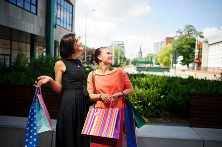 Young women with bright bags in their hands. Two cute girlfriends stand in the courtyard of the shopping center. They looking somewhere up. On the faces of the women smile.の写真素材