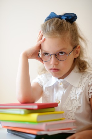 Cute little schoolgirl pondered. Girl in glasses sits leaning on a stack of books. She had a serious expression on his face.の写真素材