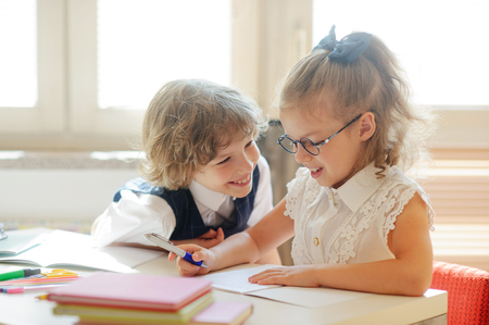 Two little classmate, laddie and girlie, are sitting at the same desk. Cute schoolgirl with glasses writes something. The neighbor on a school desk looks at the girlie. Back to school.の写真素材