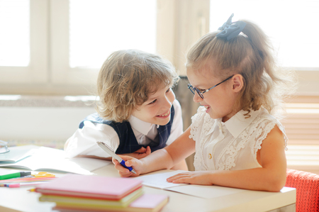 Two little classmate, laddie and girlie, are sitting at the same desk. Children learn in elementary school. Cute schoolgirl with glasses writes something. Neighbor on a school desk looks at the girl.の写真素材