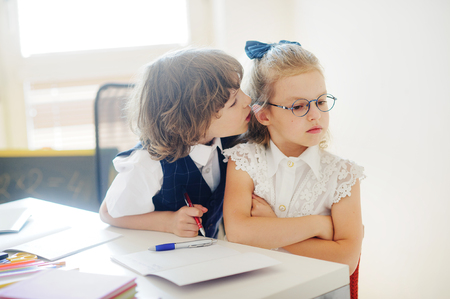 Primary school pupils sit at one desk. Cute schoolgirl angry. The boy whispers something in her ear. On a school desk children have school accessories. Back to school.の写真素材