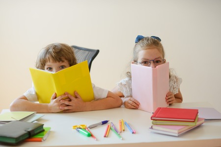 Two young classmate, boy and girl, are sitting at the same desk. Children learn in elementary school. In the hands of the students open textbooks. Kids fun look into the camera. Back to school.の写真素材