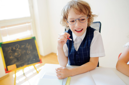 Cheerful little student bespectacled sits at school desk. It is a disciple of an elementary school. In a hand at boy ball pen. On a school desk copybooks lay. Near to a school desk a small blackboard.の写真素材