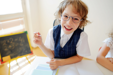 Funny little schoolboy in glasses sits at a school desk. It is a disciple of an elementary school. In a hand at boy ball pen. On a school desk copybooks lay. Near to a school desk a small blackboard.の写真素材
