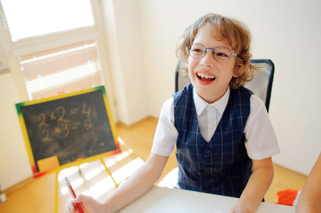 Cheerful little student bespectacled sits at school desk. It is a disciple of an elementary school. In a hand at boy ball pen. On a school desk copybooks lay. Near to a school desk a small blackboard.の写真素材