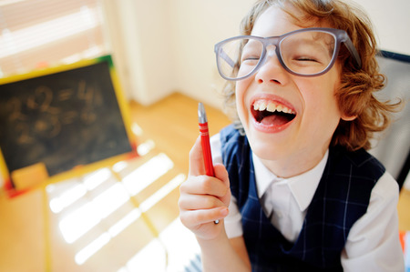 Cheerful little student bespectacled sits at school desk. It is a disciple of an elementary school. In a hand at boy ball pen. On a school desk copybooks lay. Near to a school desk a small blackboard.の写真素材