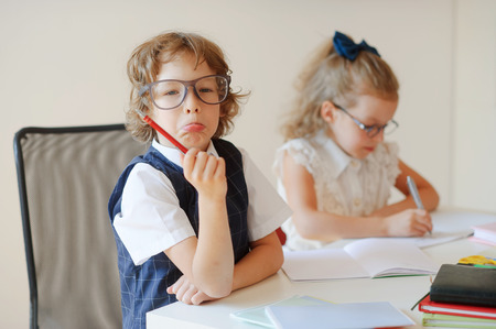 Funny little pupils sit at one desk. They are pupils of an elementary school. Boy fools around, girlie writes something with a serious look. On a school desk children have school accessories and textbooks.の写真素材