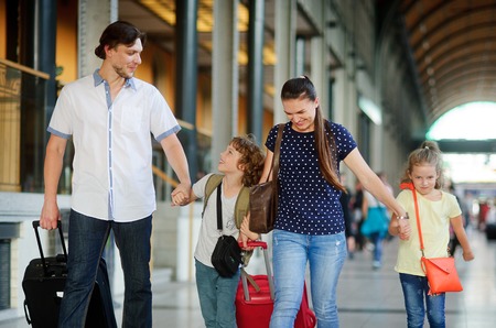 Young family with two children at train station. The family passes through the big waiting room. All hold hands. The father and the mother have big suitcases. In the hall many passengers.の写真素材